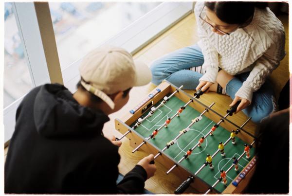 Image of two teens playing foosball on a floor next to a window.