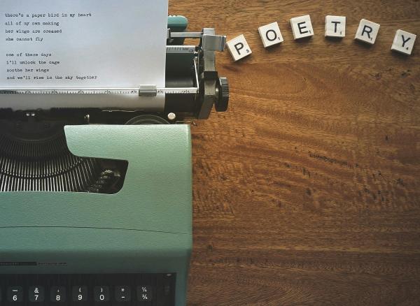 A green typewriter on a wooden desk writing poetry. There are scrabble tiles spelling "Poetry" beside it.