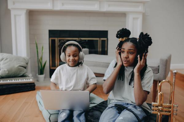Two children listening, with headphones, to something on a computer. A trumpet sits next to them in a living room.