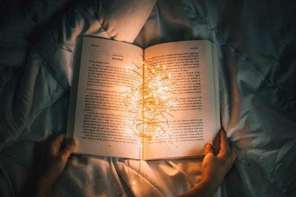 An open book held on a blanket with a string of lights illuminating it