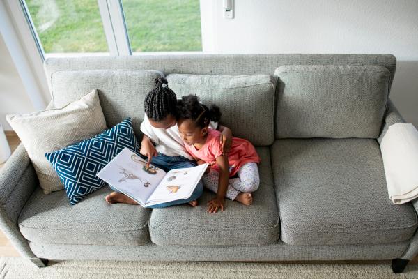 two Black girls sit on a grey couch reading a storybook together; above view