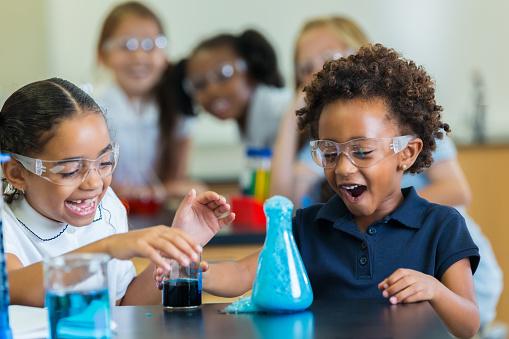 two Black elementary-school girls laugh at chemistry experiment with blue foam in beaker