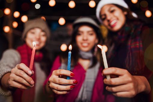 unfocused photograph of three young women in hats and scarves, each holding a candle close to the camera