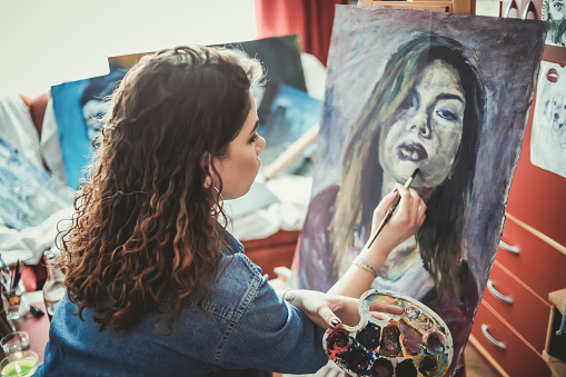 long-haired brunette artist paints a self-portrait in grays and dark colors