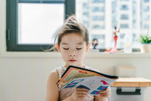 young Asian girl reads a magazine in brightly lit room with window behind her